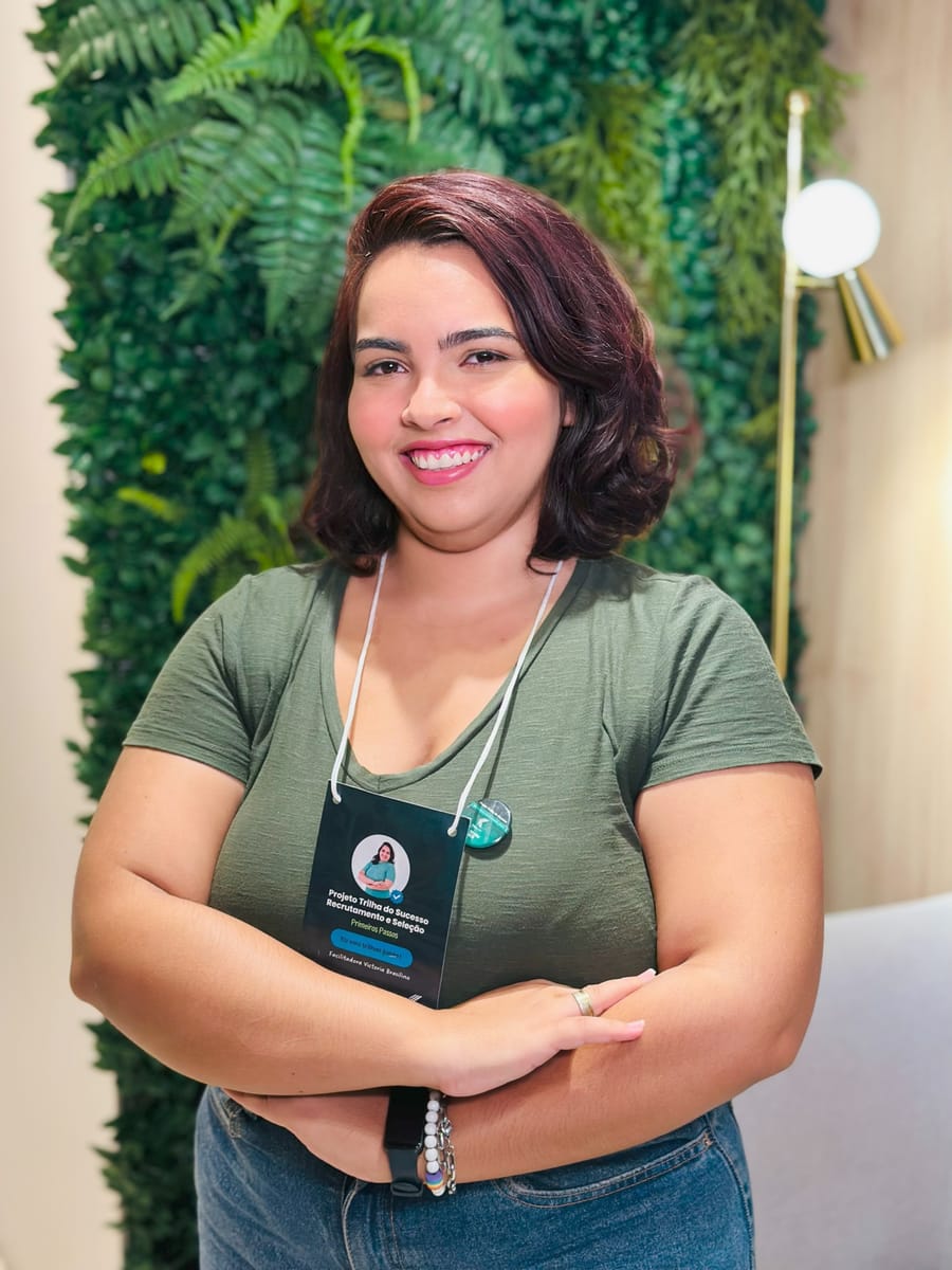 Victoria de braços cruzados e crachá de evento, em frente a uma parede verde