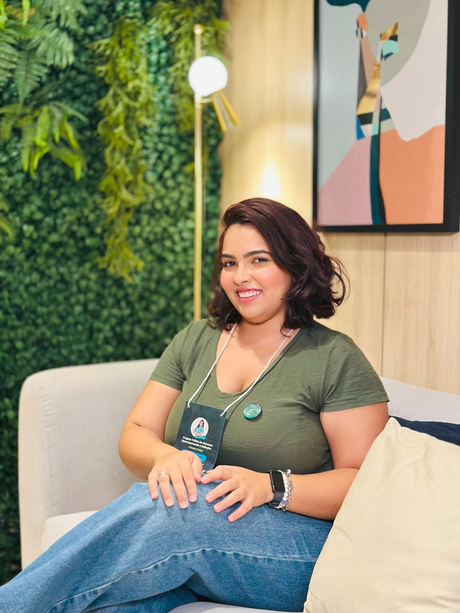 Victoria sentada num sofá claro diante de uma parede de plantas, com crachá de evento, sorrindo