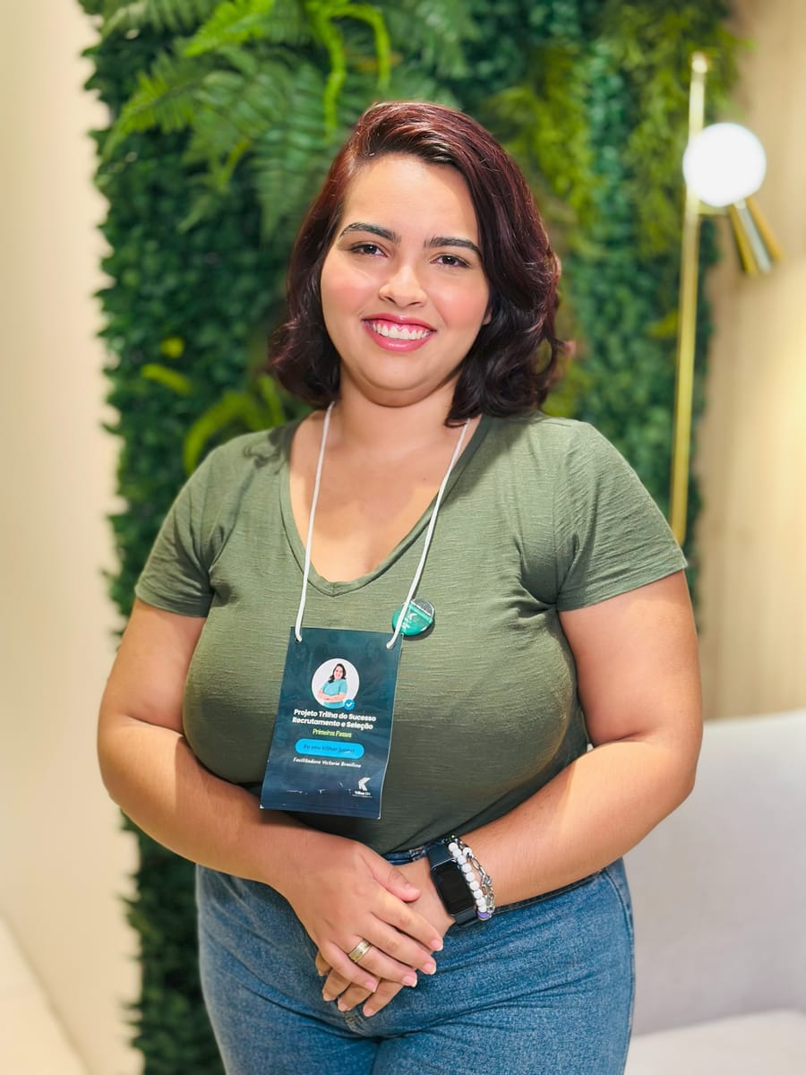 Victoria sorrindo, com as mãos unidas e crachá de evento, ao lado de uma luminária dourada
