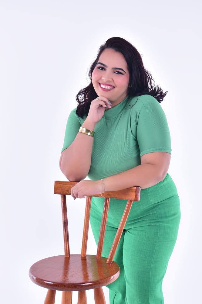 Victoria sorrindo, apoiada no encosto de uma cadeira de madeira, com a mão no queixo