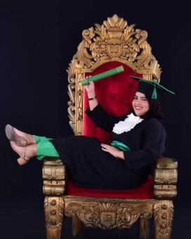 Victoria de beca preta e capelo verde, sentada num trono dourado de veludo vermelho, erguendo o canudo
